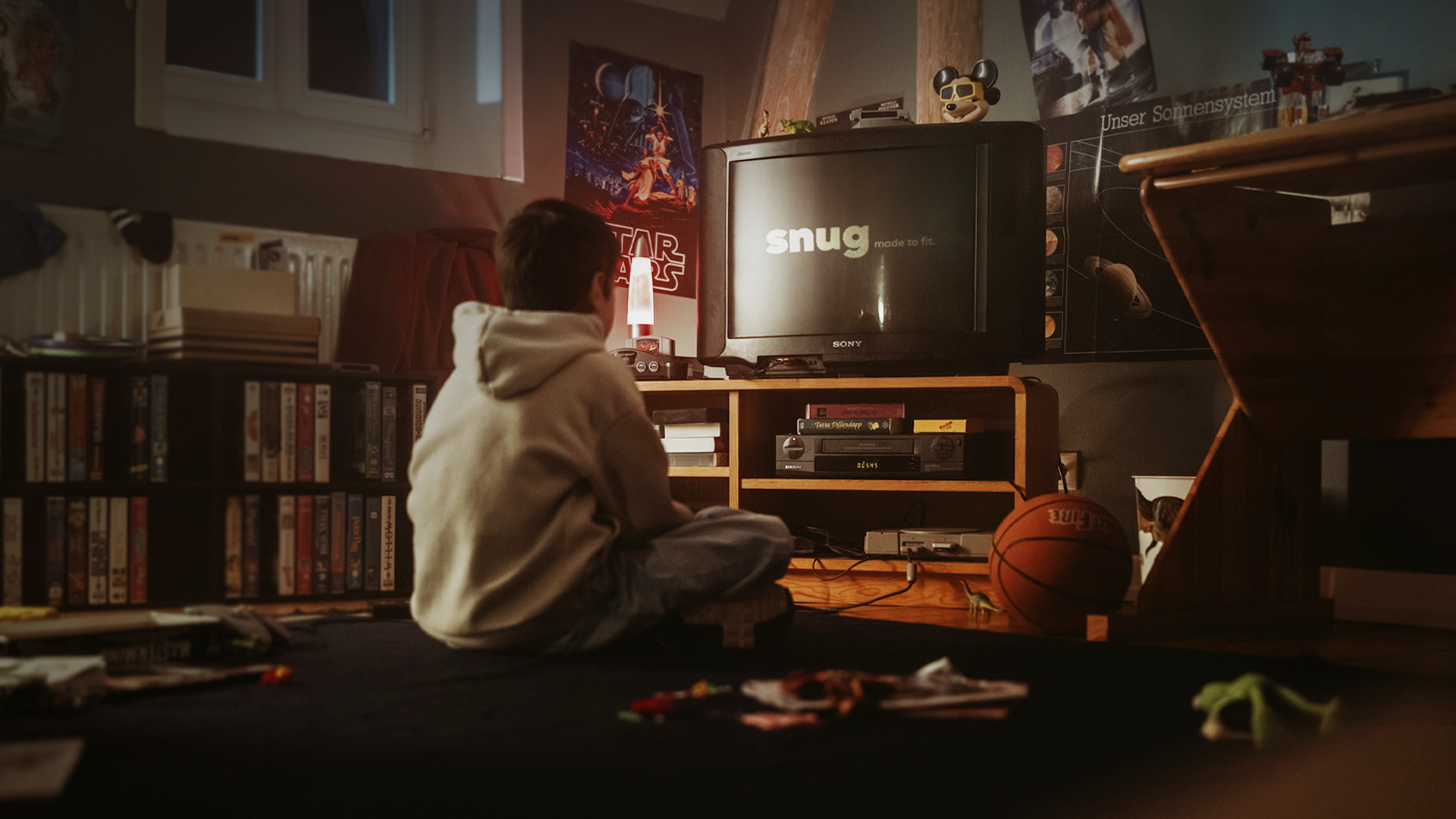 A boy sits with legs crossed in a lamplit 90s bedroom, watching a CRT TV that reads snug, made to fit, a still from snug's own launch campaign.